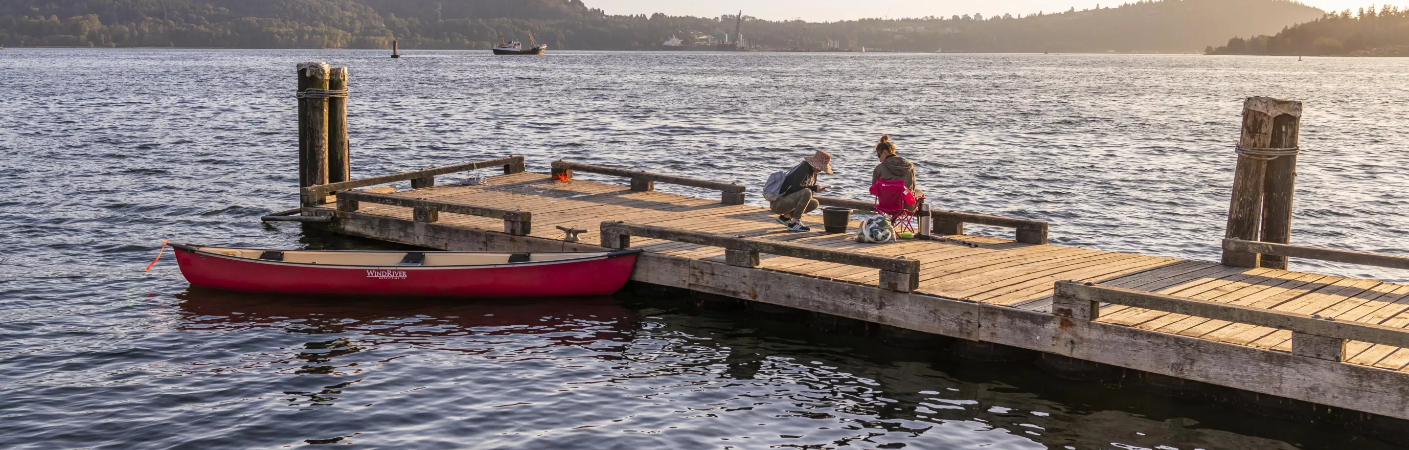 A red canoe sits in the water, tied to a dock. Two people sit on the dock to the right of the canoe..