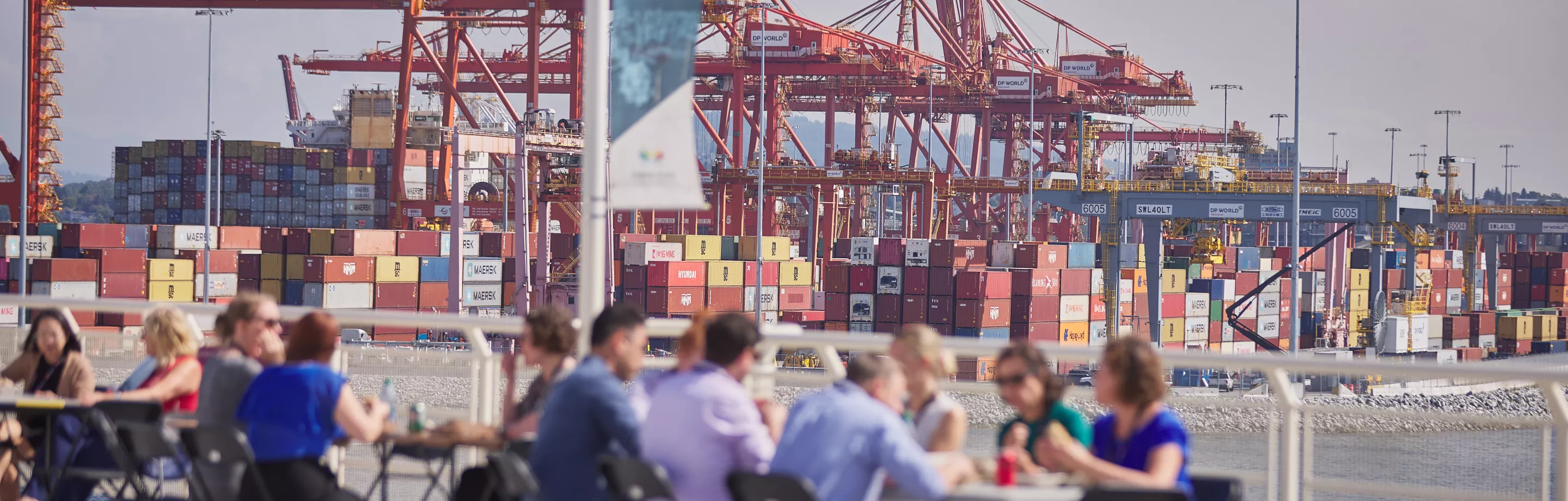 Port authority employees sit at picnic tables outdoors. Orange gantry cranes and shipping containers can be seen in the background.