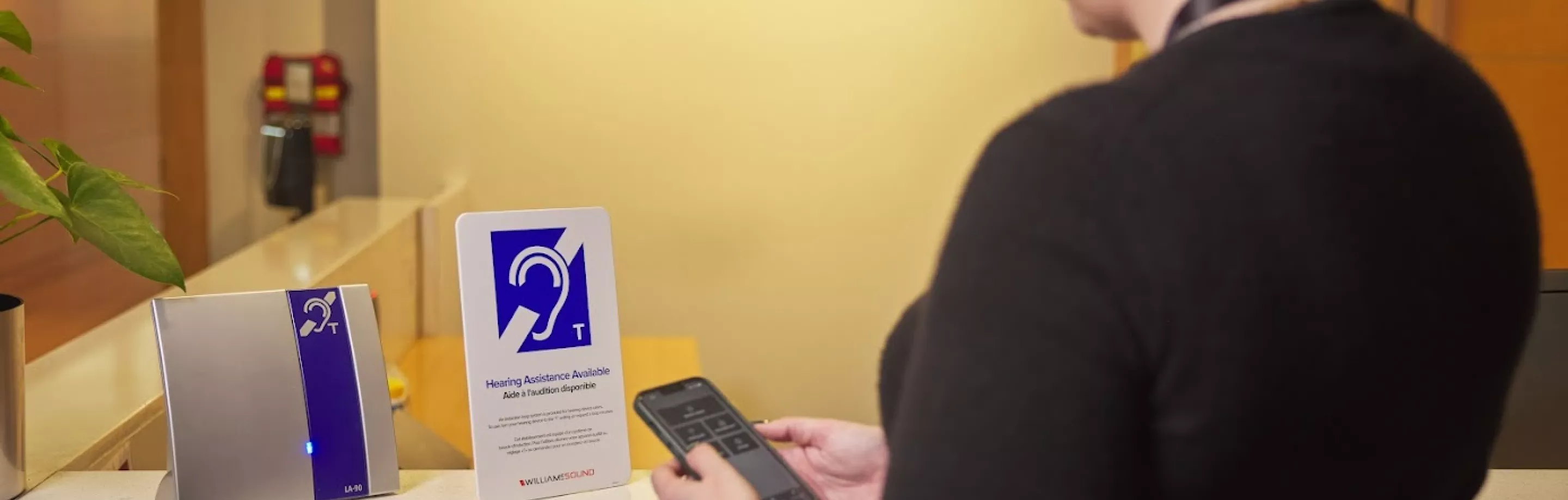 A woman in a dark sweater stands at a counter looking at a hearing loop sign