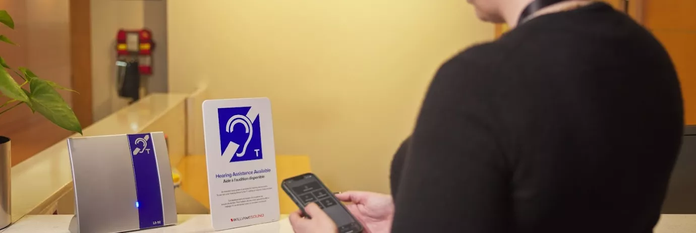 A woman in a dark sweater stands at a counter looking at a hearing loop sign