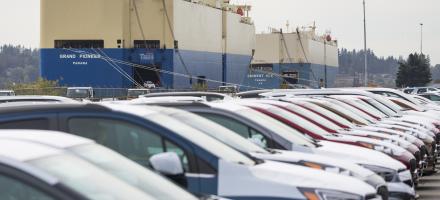 A row of cars in a lot. A ro-ro ship can be seen in the background.